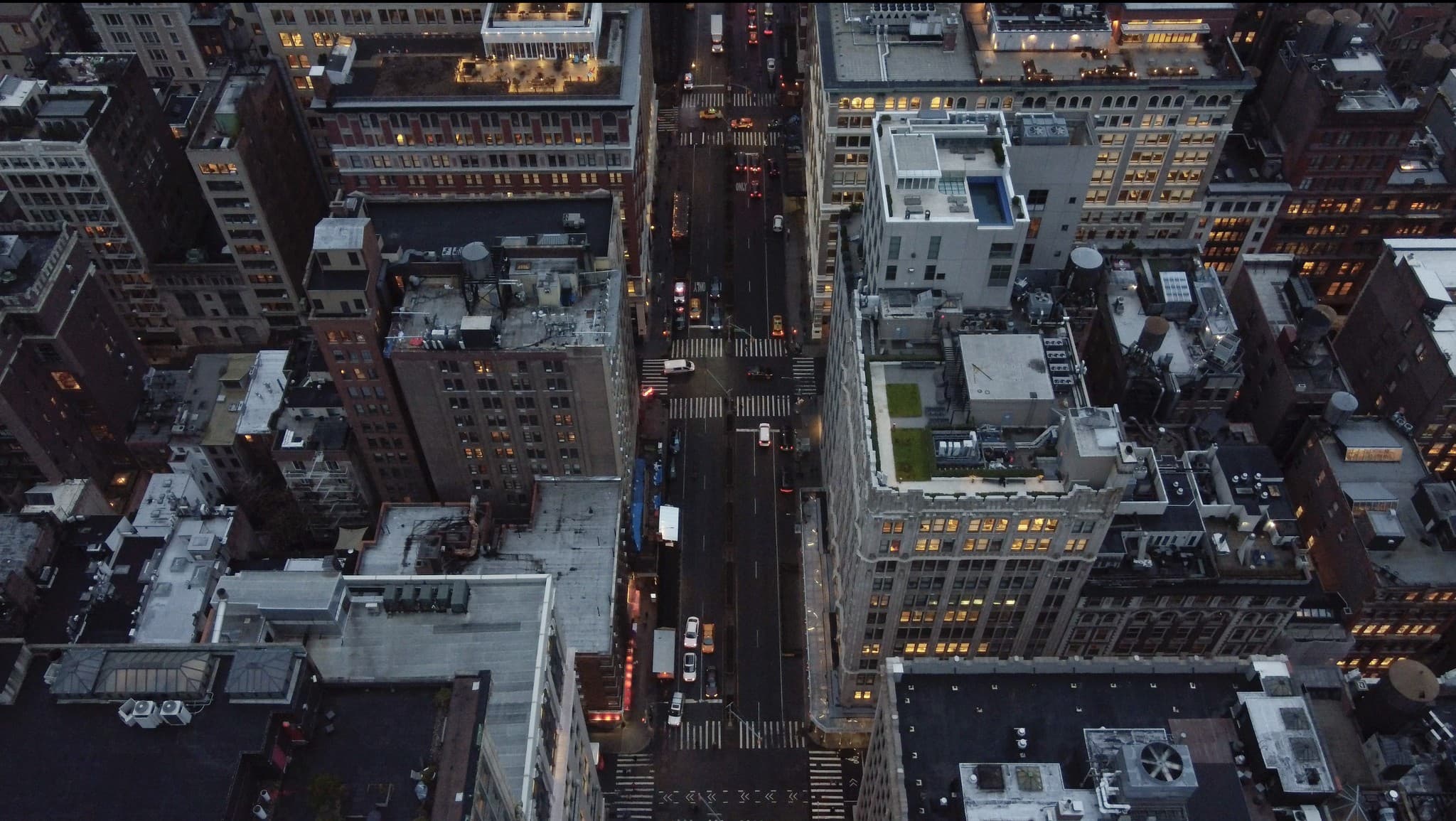 Top-down aerial view of a Manhattan intersection at dusk, rooftops and lit windows below